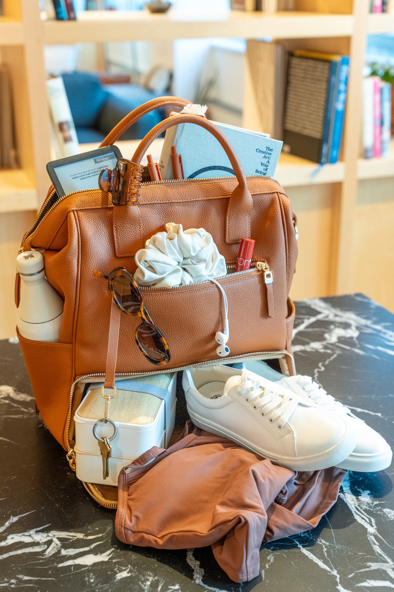 Brown tan leather work backpack with items on a marble surface, with a bookshelf in the background